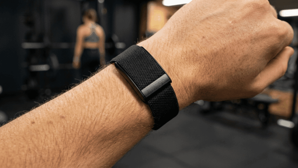 close-up of a person wearing a Whoop fitness band on wrist in a gym setting, highlighting its sleek, screen-free design and strap fit
