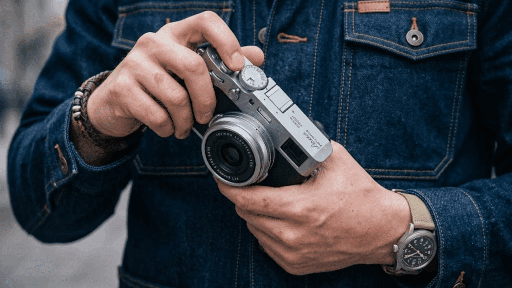 person holding a Fujifilm X100VI camera and adjusting the top dial while wearing a denim jacket and wristwatch outdoors.