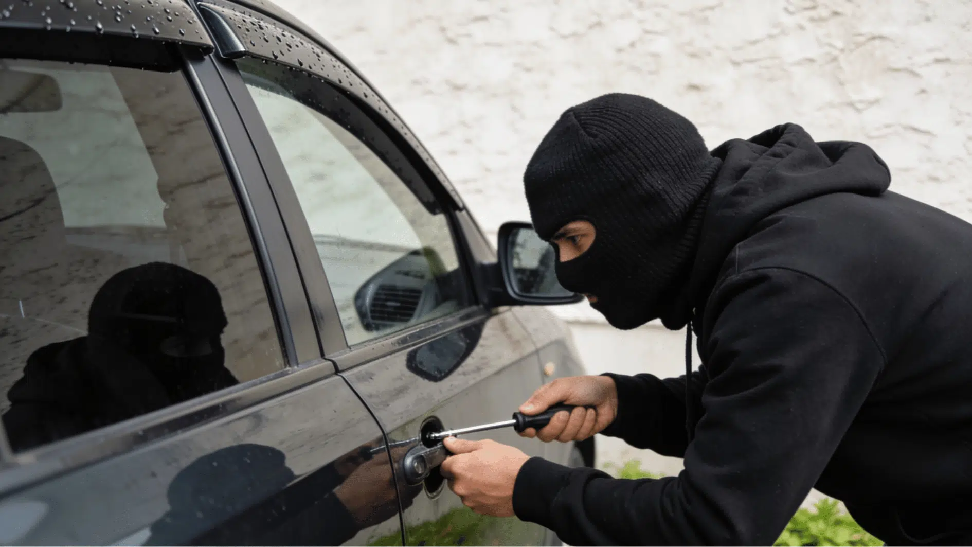 masked person attempting to break into a car door using screwdriver outside building realistic