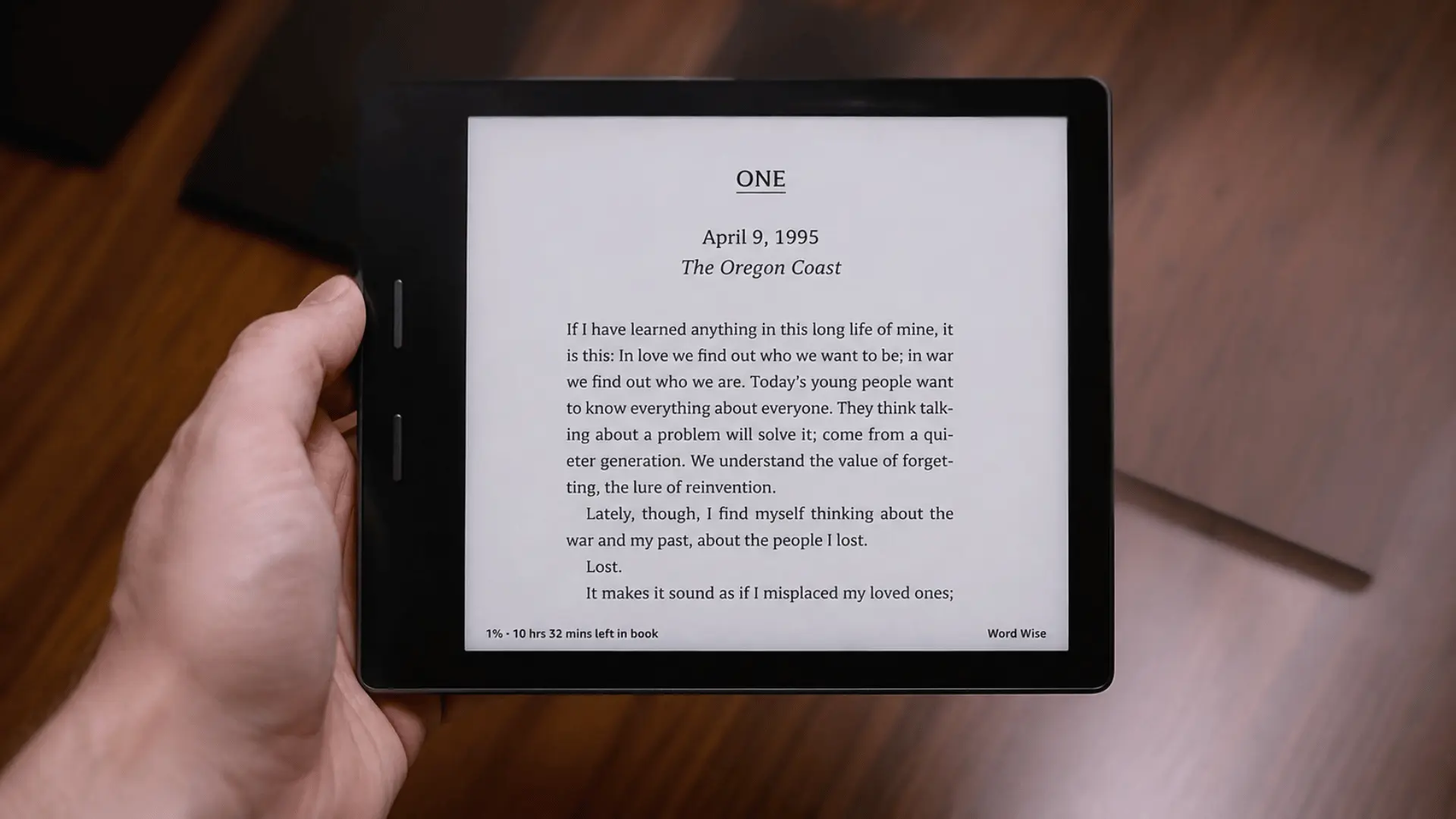 Black e-reader held in one hand displaying a chapter opening, with wooden table in the background