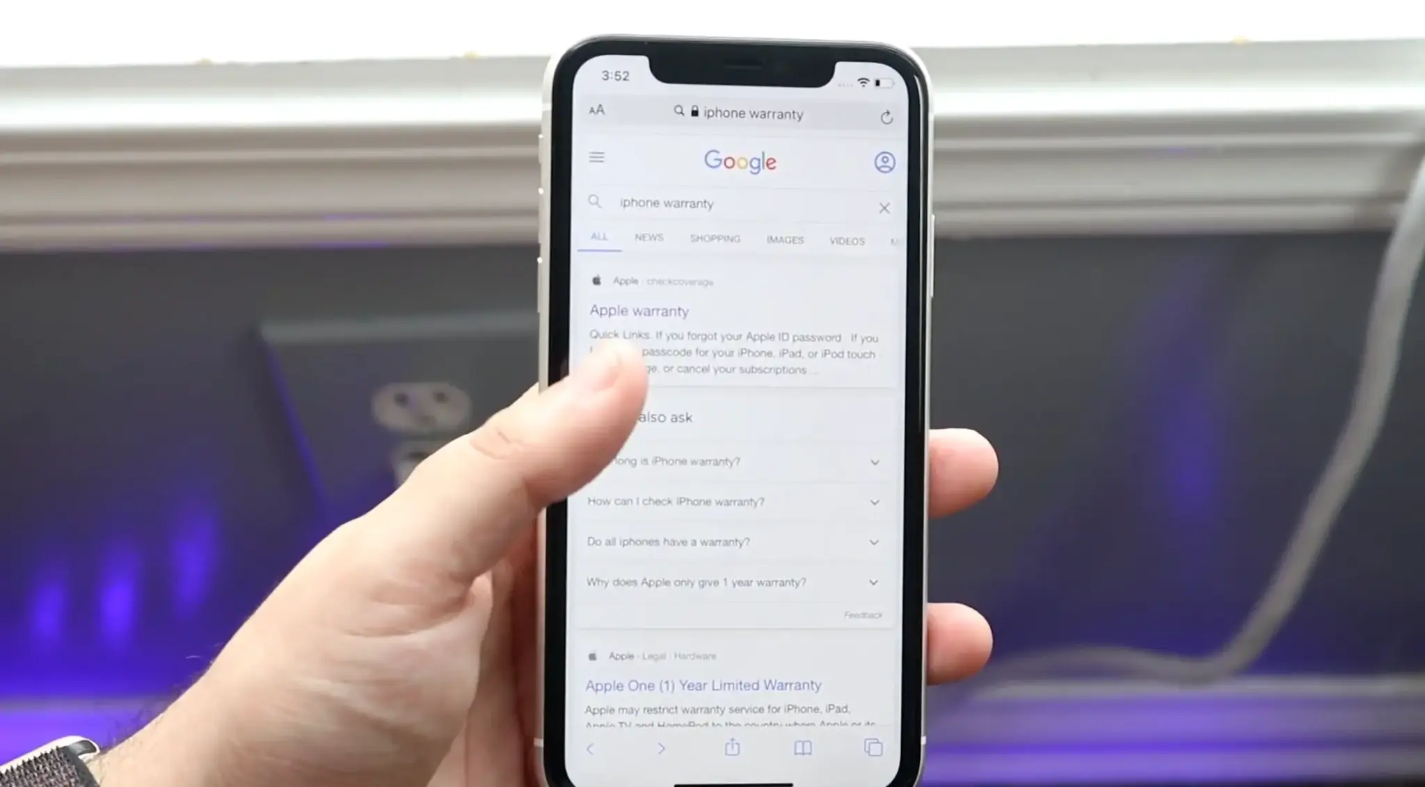 Hand holding an iPhone displaying search results for Apple’s iPhone warranty and coverage page.