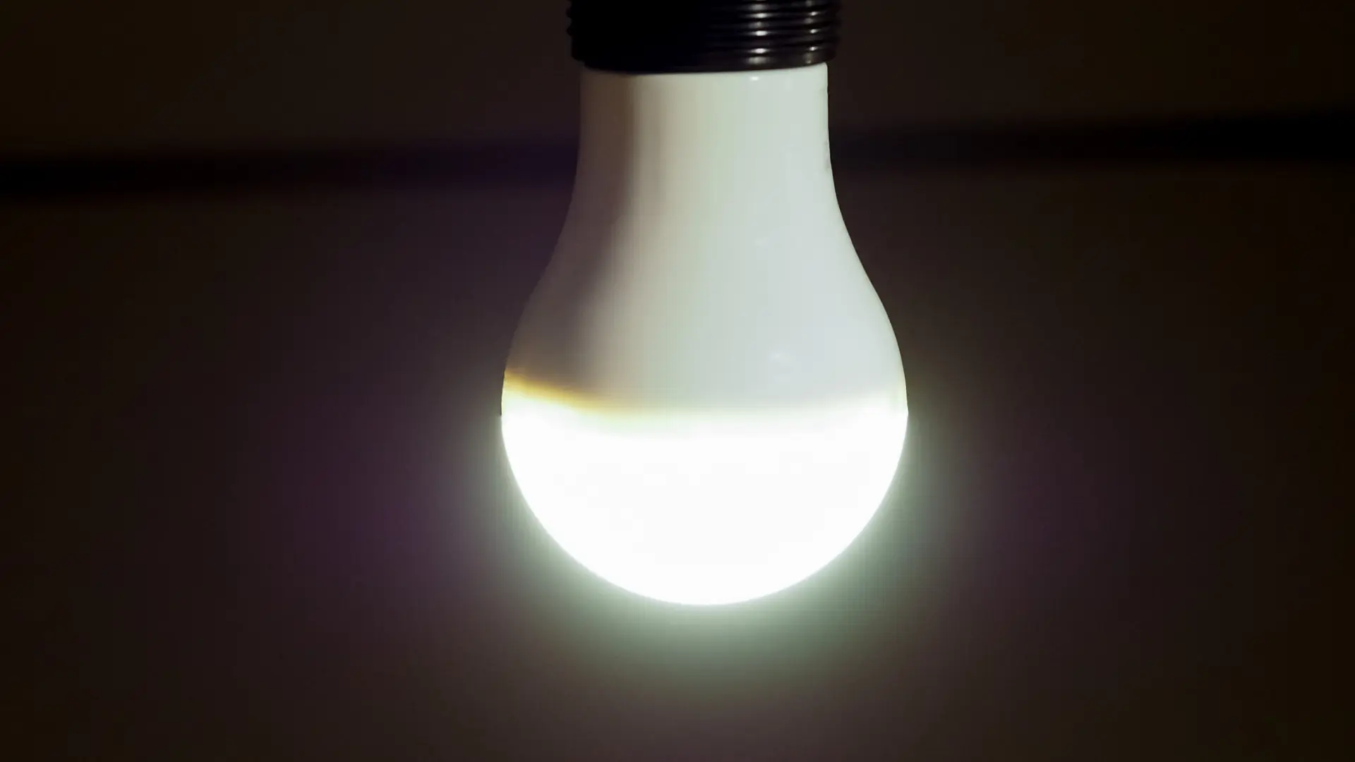 Close-up of a glowing white LED light bulb hanging against a dark background, illuminating the lower half with bright light