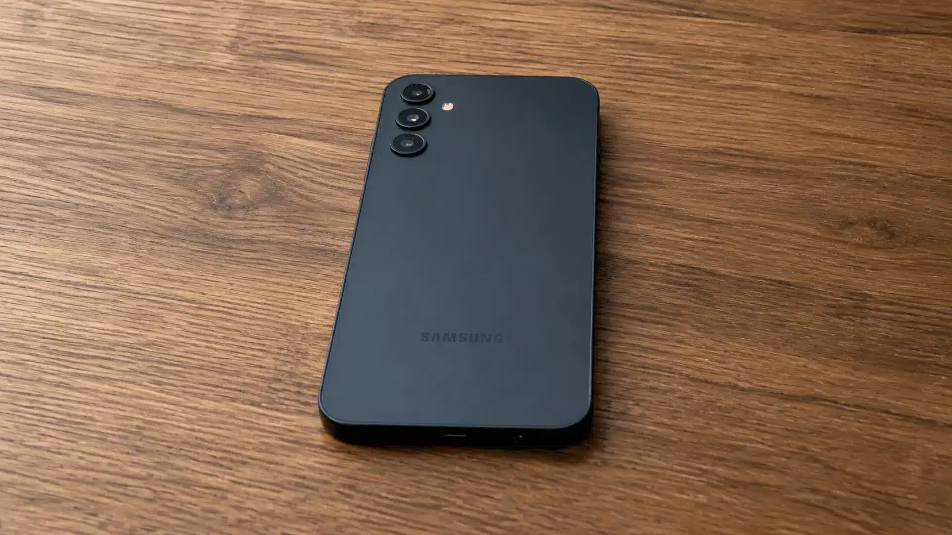 Black Samsung smartphone with triple rear cameras resting on a warm wooden surface.