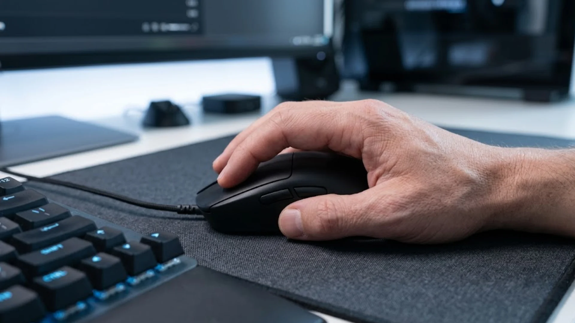 Hand resting on a black wired gaming mouse beside a blue-lit mechanical keyboard