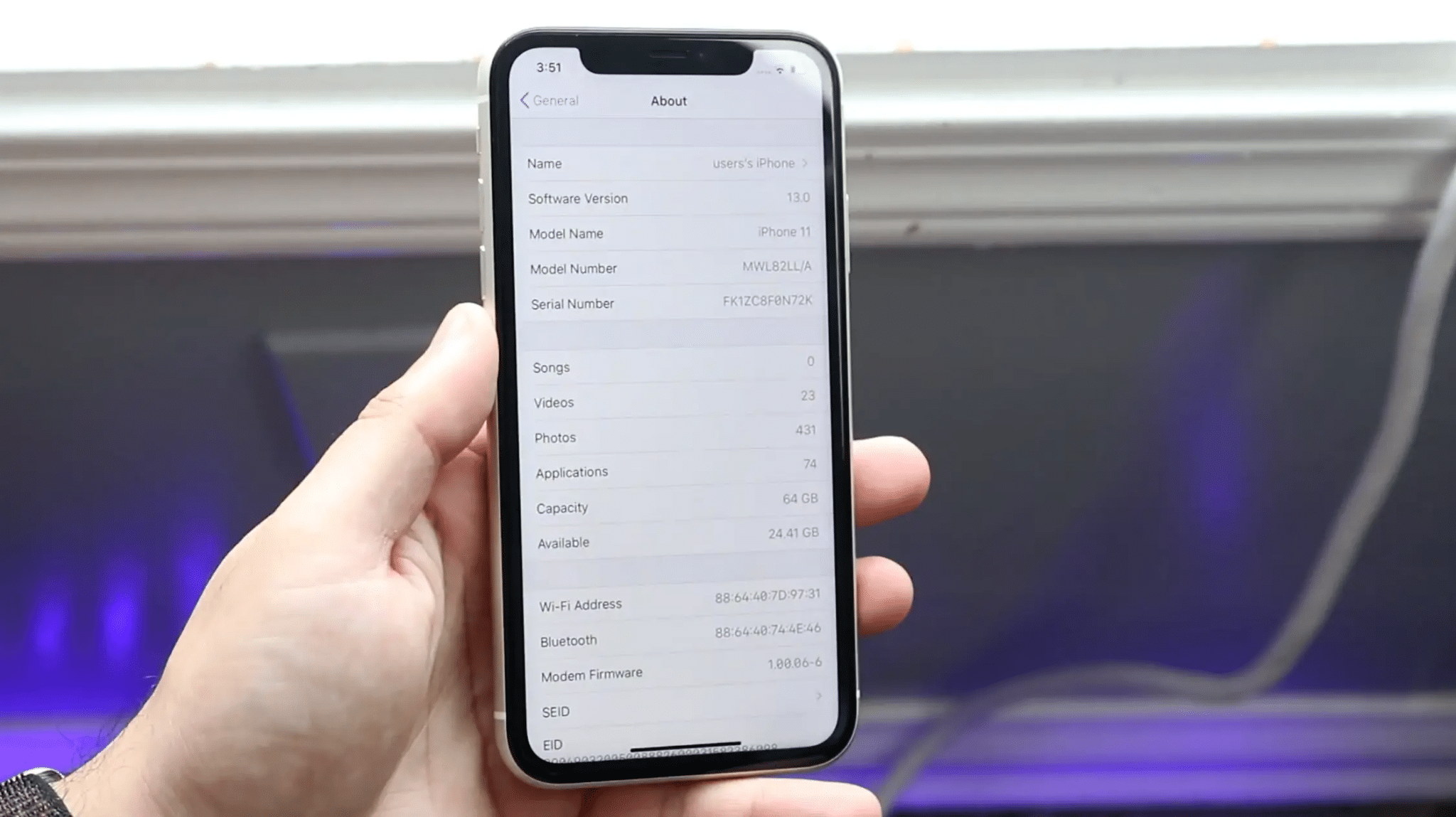 Hand holding an iPhone with the About screen showing the model name, model number, serial number, and storage details.