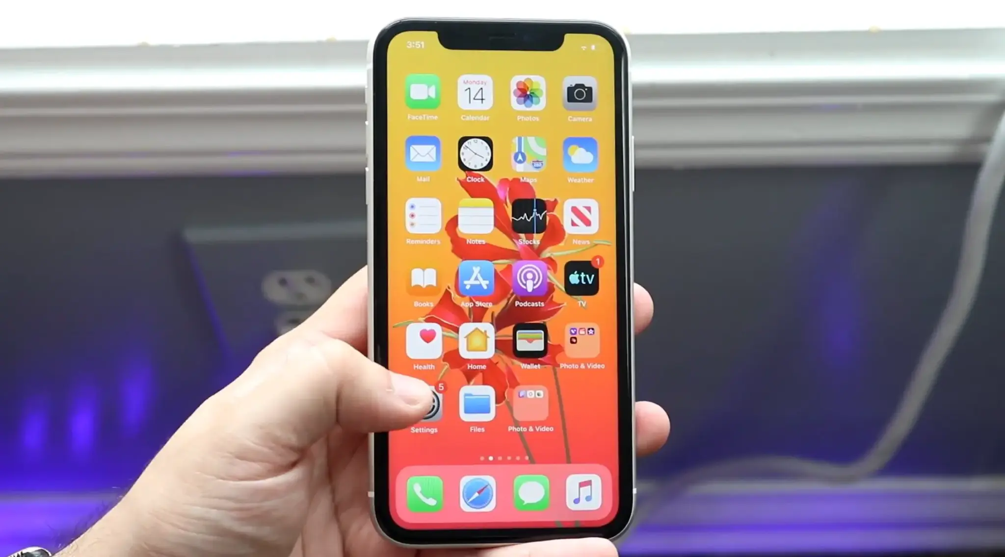 Hand holding an iPhone with a thumb pointing toward the Settings app on the Home Screen.