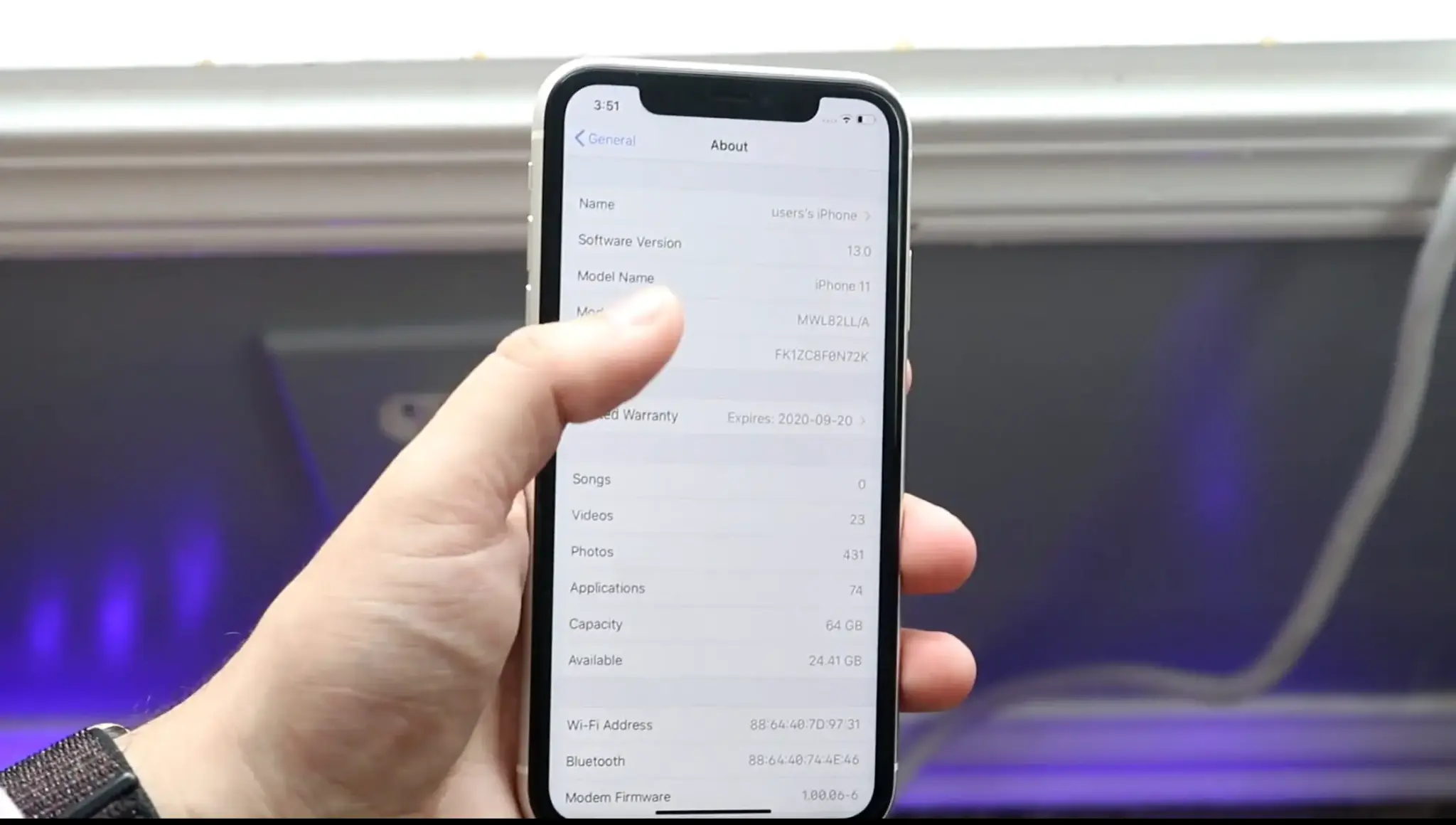 Hand holding an iPhone with a thumb pointing to the iPhone 11 model name on the About screen.