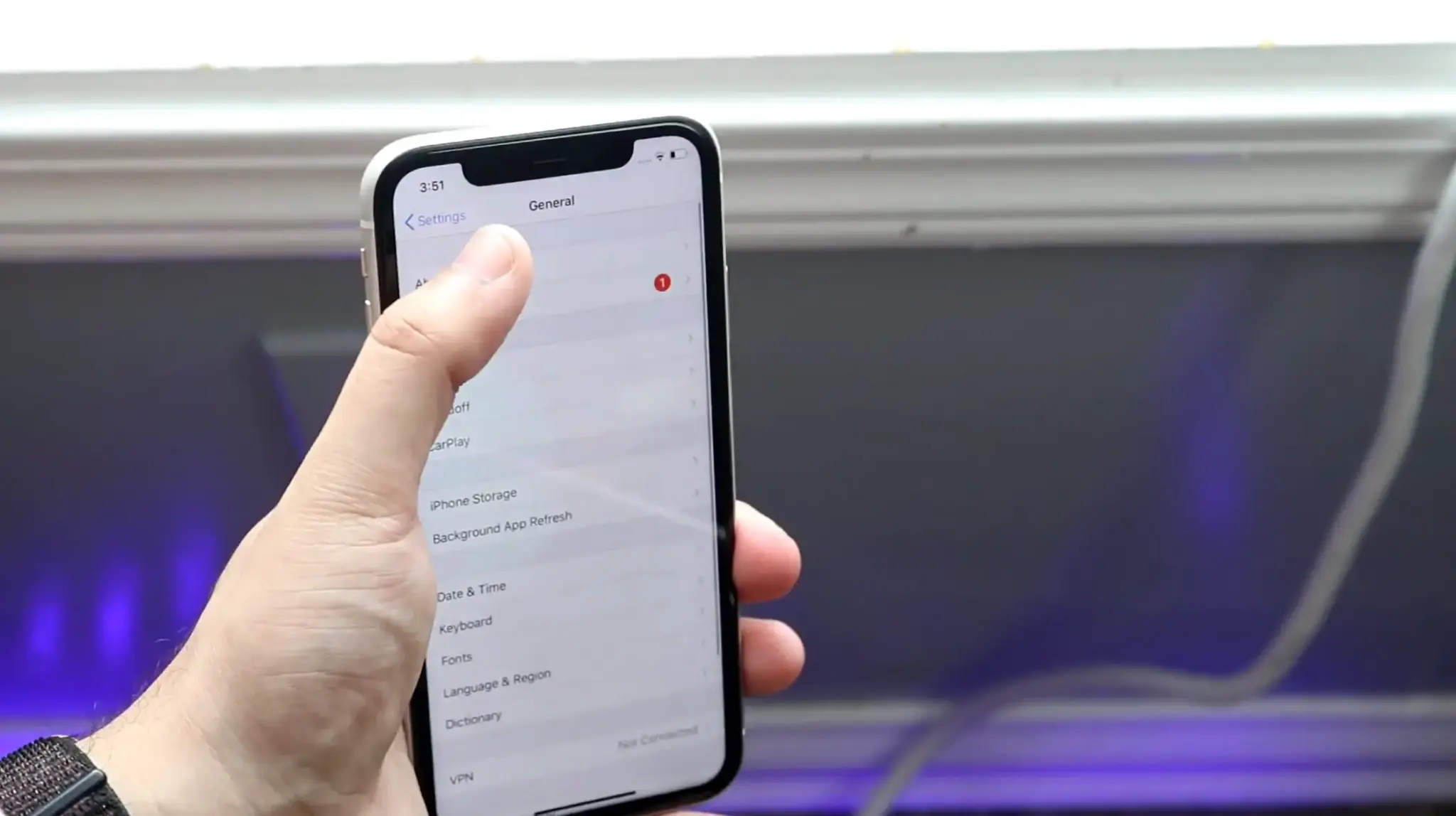 Hand holding an iPhone with the General settings menu open and a thumb pointing toward About.
