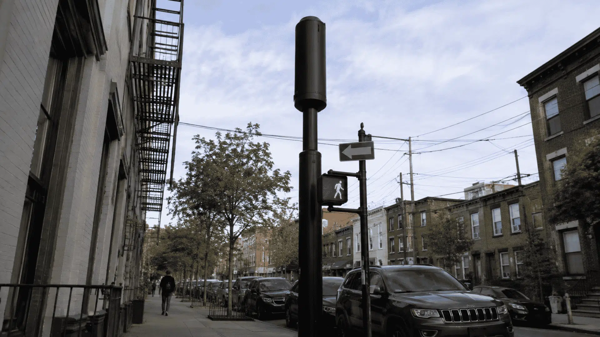 black small cell tower on city street beside brick buildings parked cars trees and overhead utility wires area